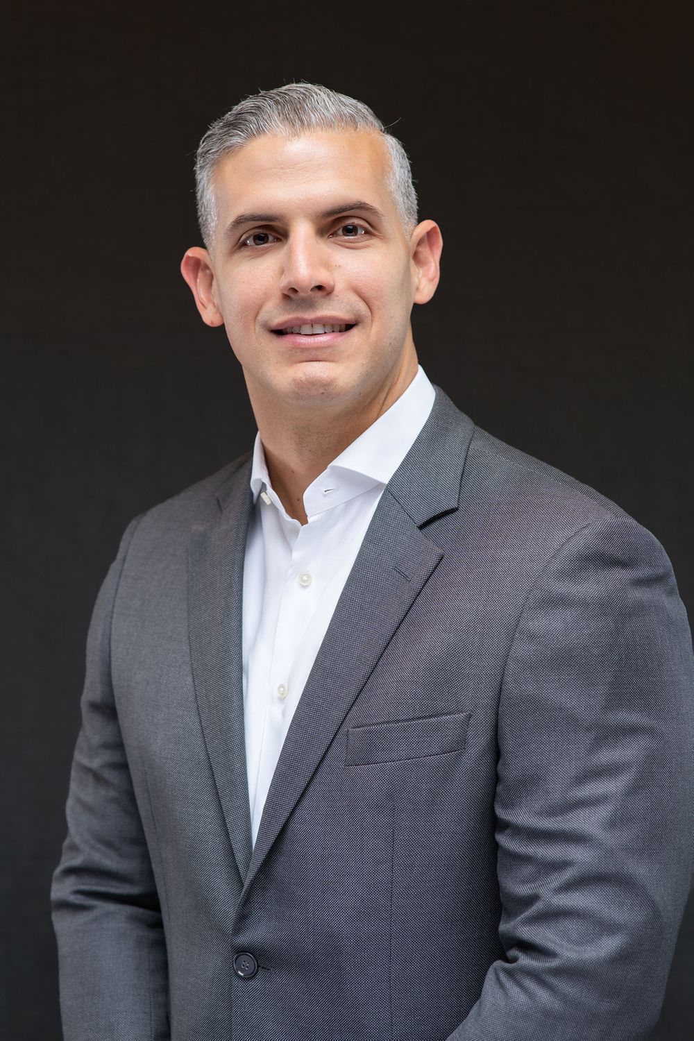 Professional headshot of a smiling man in a gray suit and white shirt against a dark background.