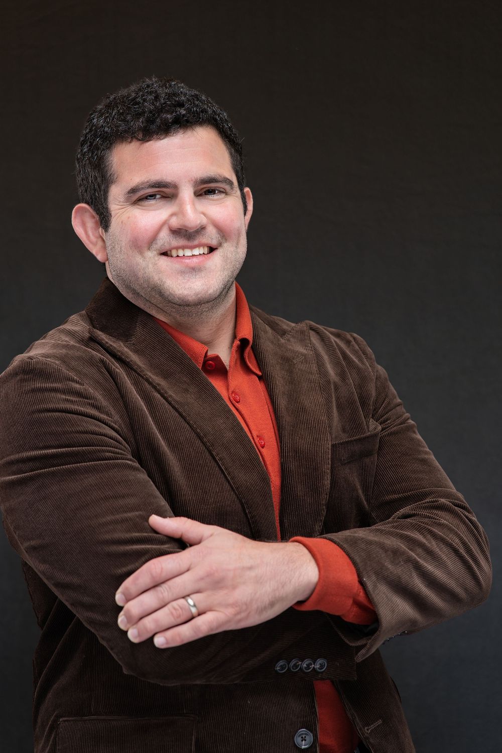 Smiling man in brown velvet blazer and red shirt with arms crossed against dark background.
