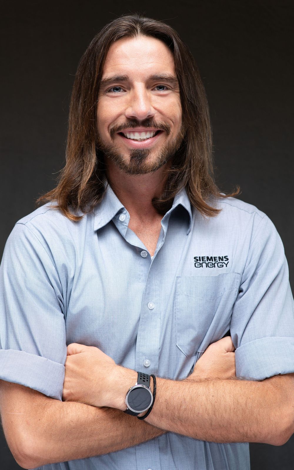 Man with long hair smiling in Siemens Energy shirt, arms crossed, professional headshot.