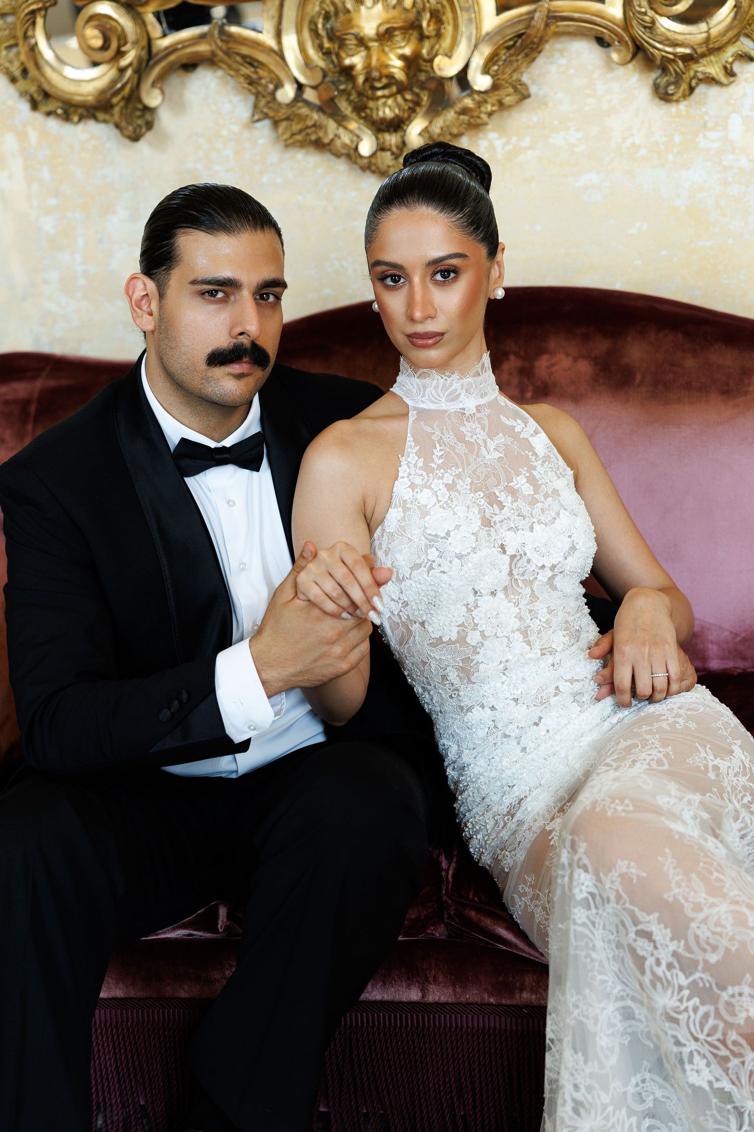 Elegant couple in formal attire sitting on velvet sofa, man in tuxedo and woman in white lace gown.
