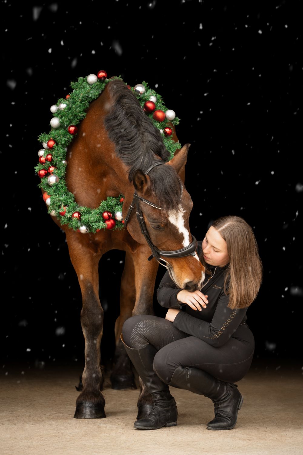 Young woman in black outfit crouches beside bay horse wearing Christmas wreath, sharing tender moment in snowy night setting.