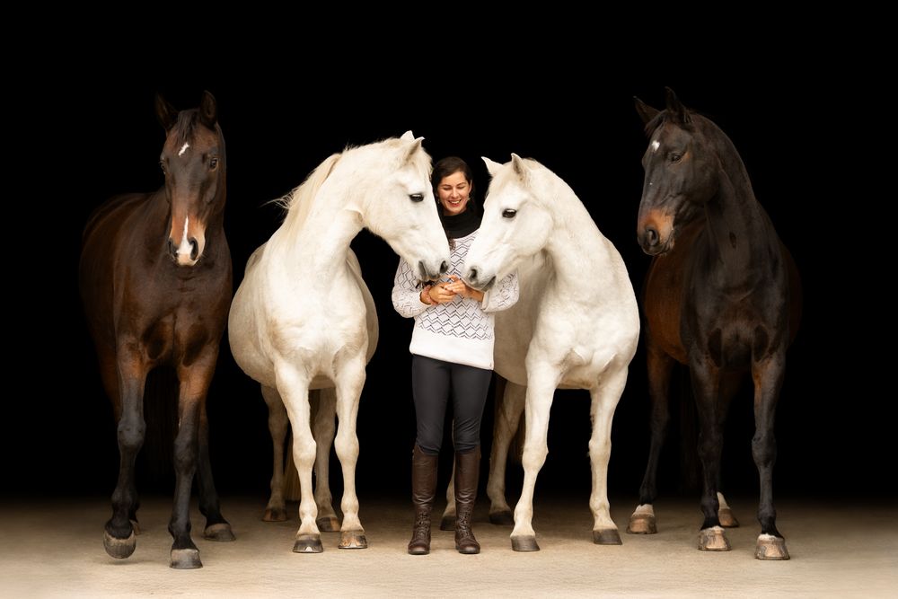 Woman standing between four horses, two white and two dark, against a black background in a studio setting.
