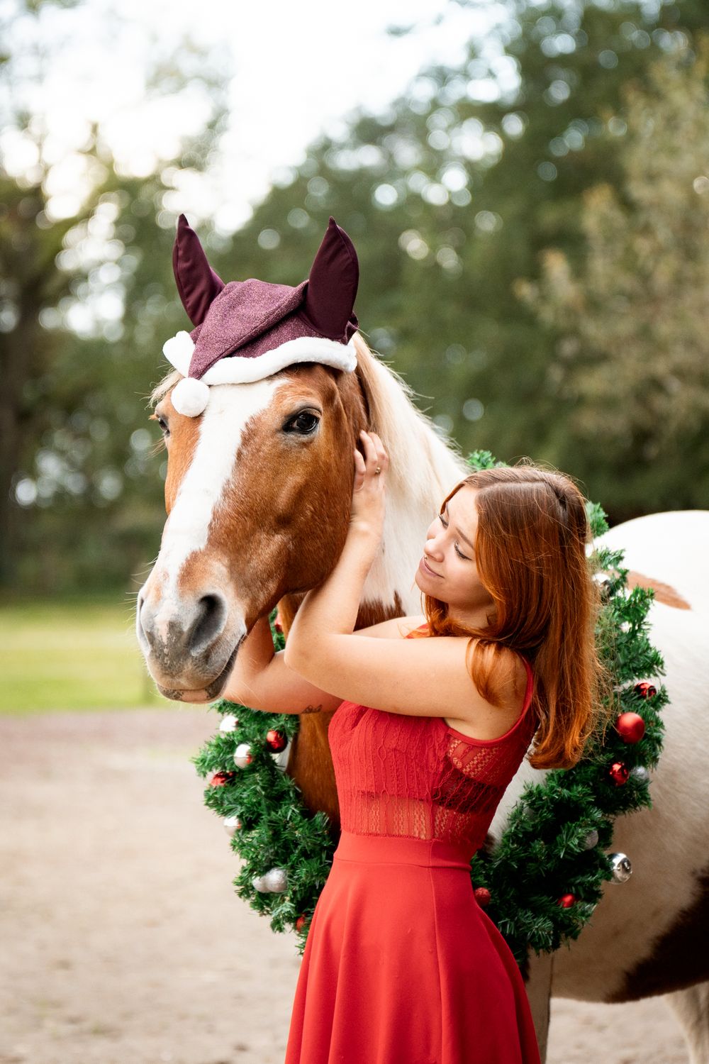 Woman in red dress hugging a horse wearing a Santa hat and Christmas wreath outdoors.