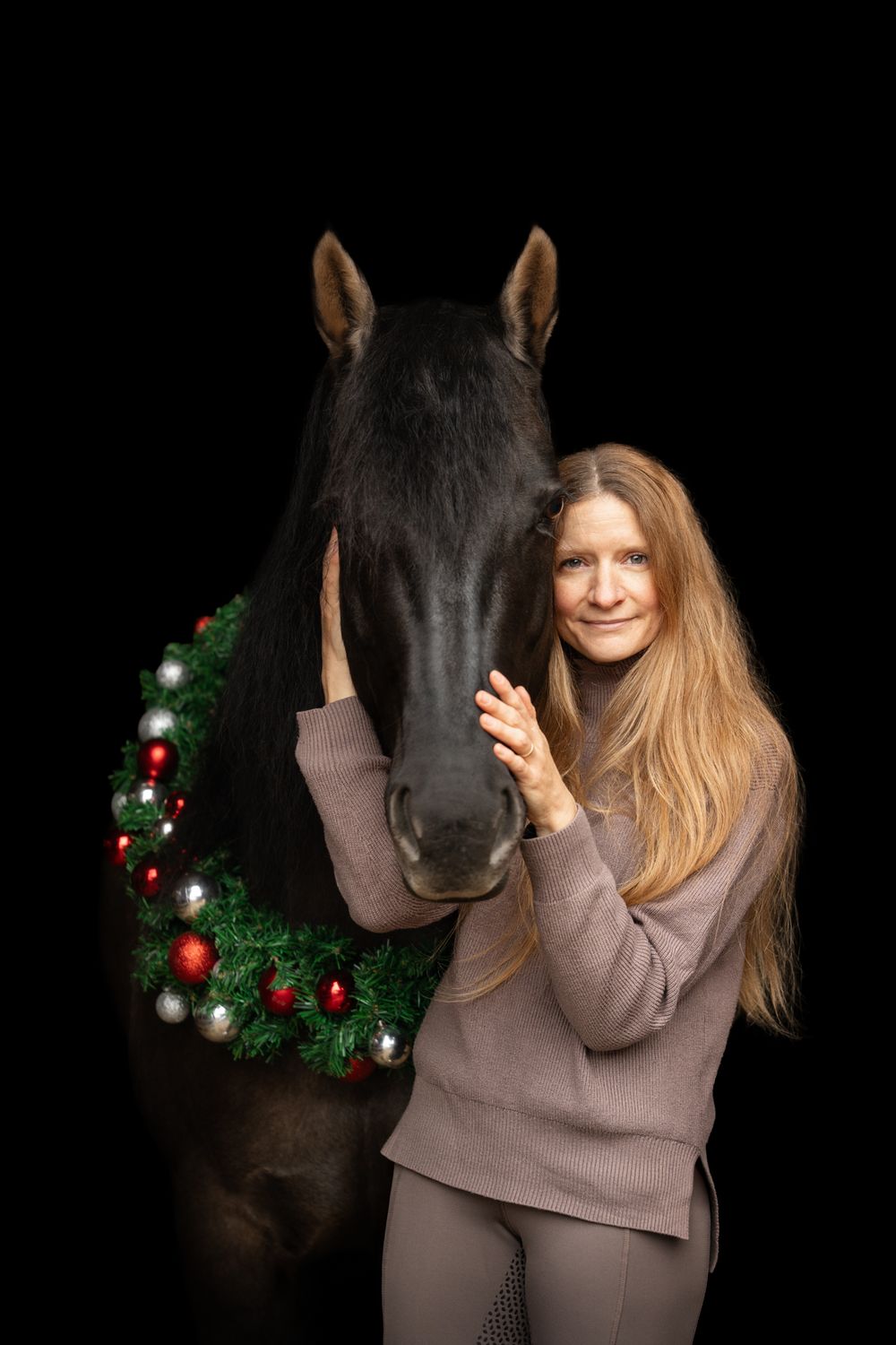 Woman with long blonde hair smiling beside a dark horse wearing a festive Christmas wreath on black background.