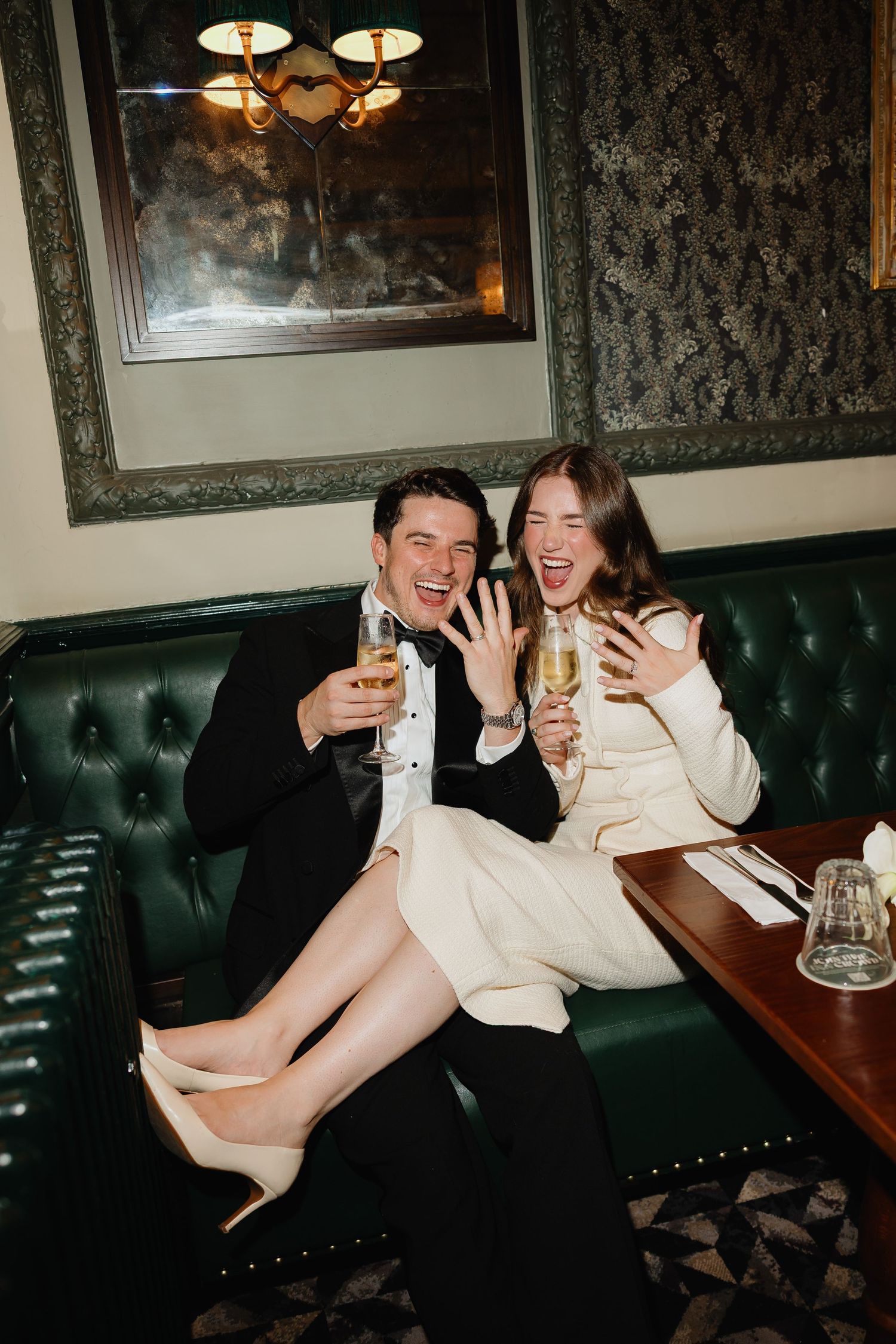 Joyful couple celebrating engagement on green leather booth, holding champagne glasses and showing off ring in elegant restaurant.