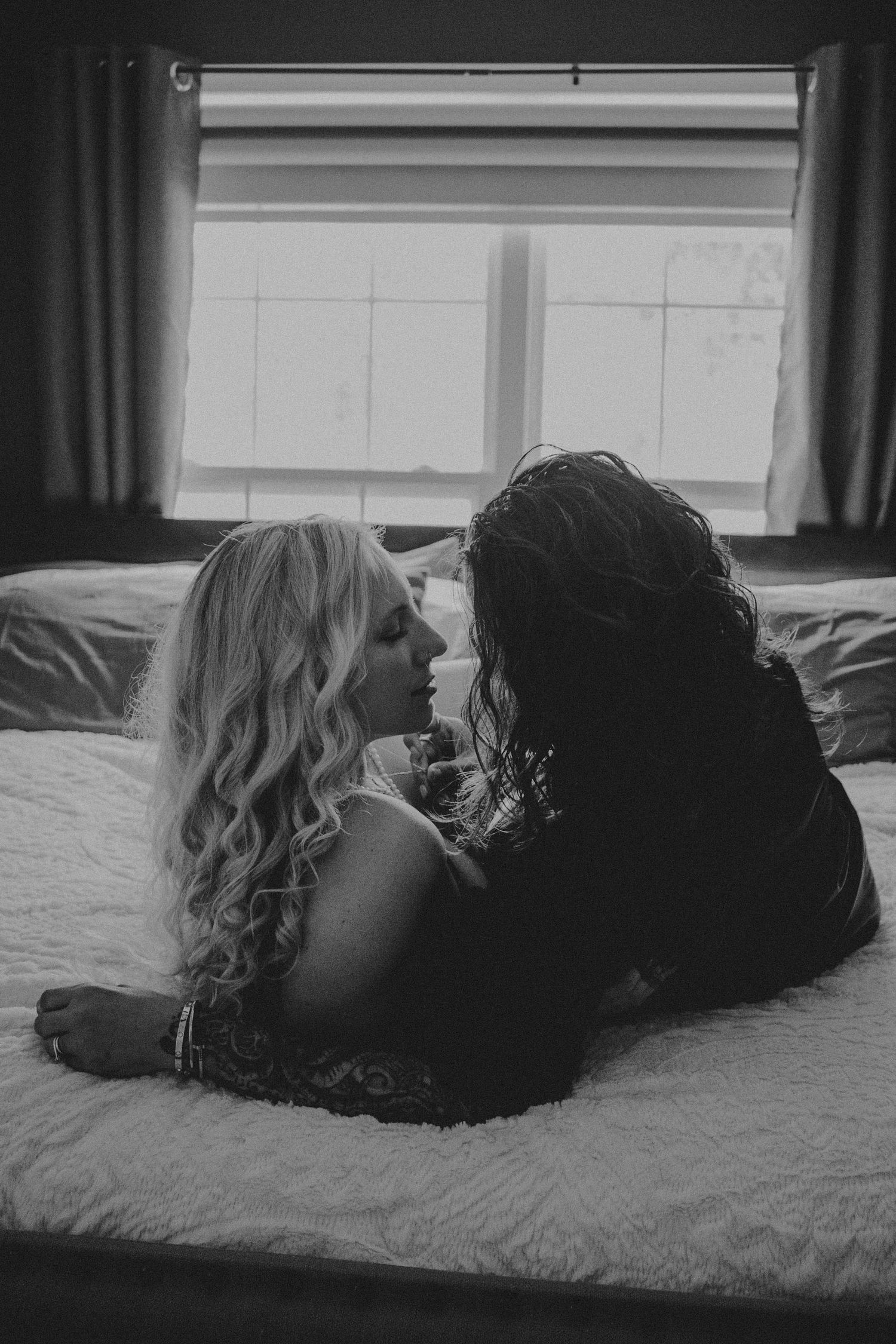 Black and white photo of two women lying on a bed facing each other near a window.