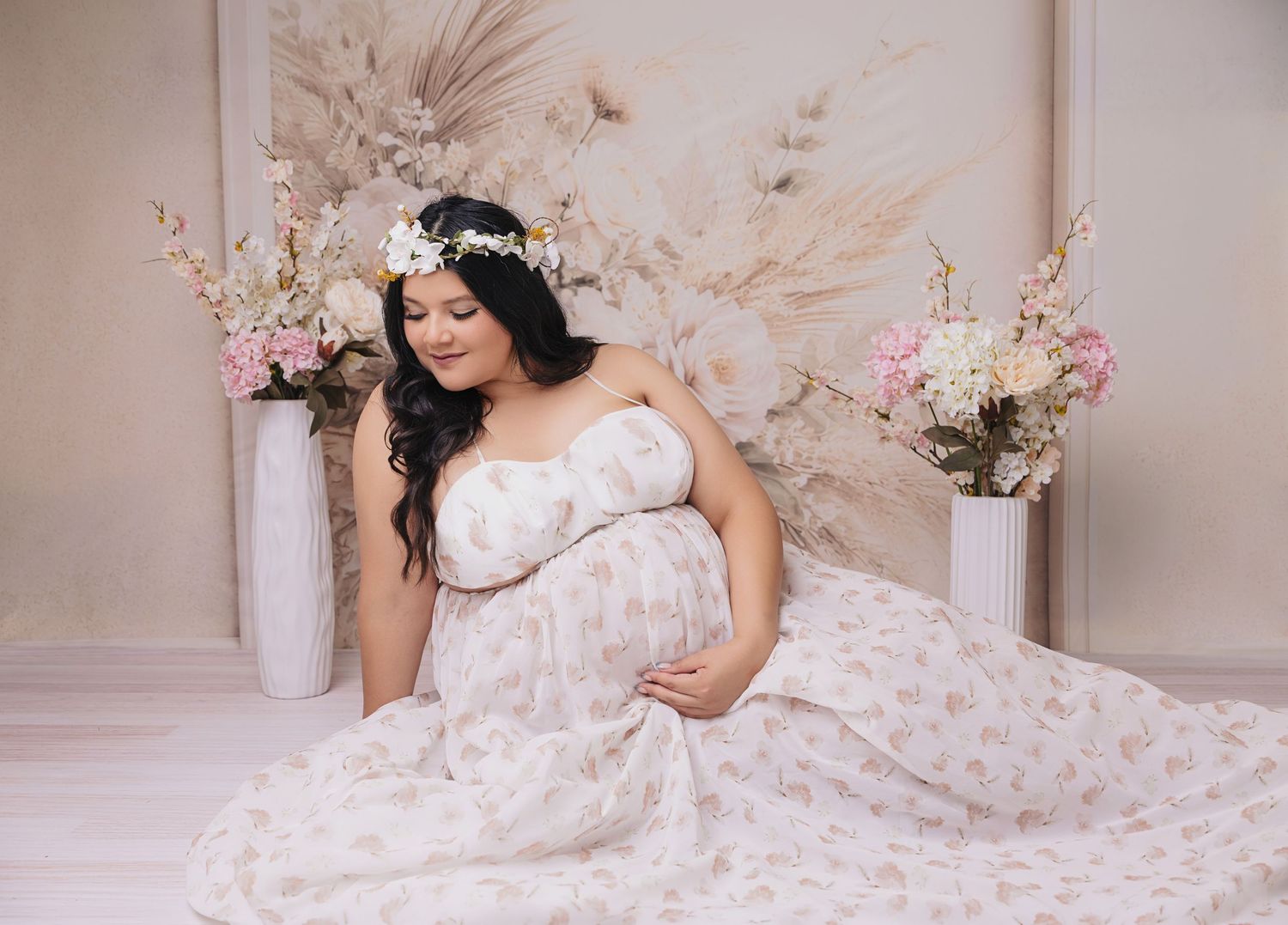 Pregnant woman in floral gown with flower crown poses gracefully among pampas grass and pink blooms in studio.