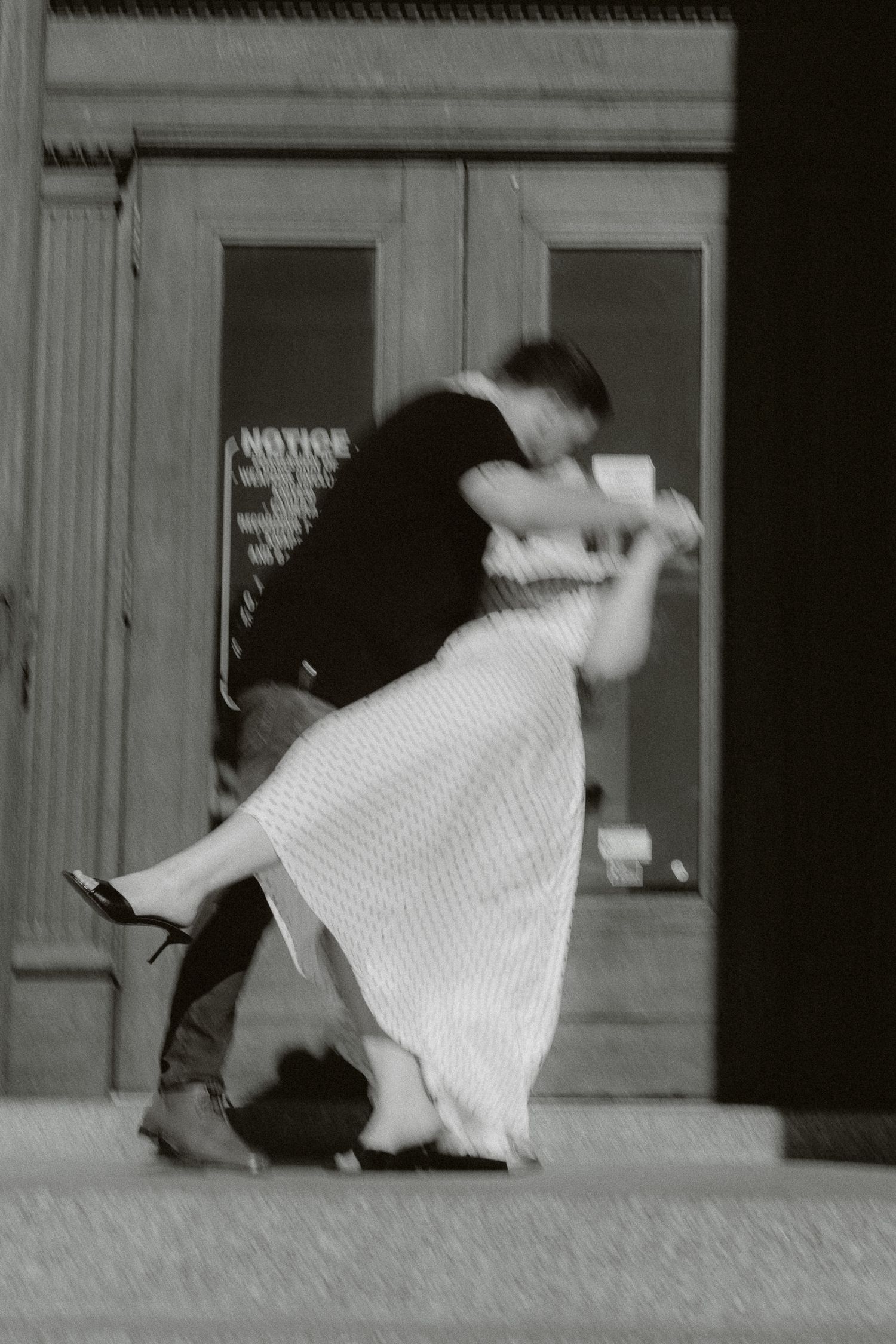 Black and white photo of a couple dancing romantically on a sidewalk in front of a building with glass doors.
