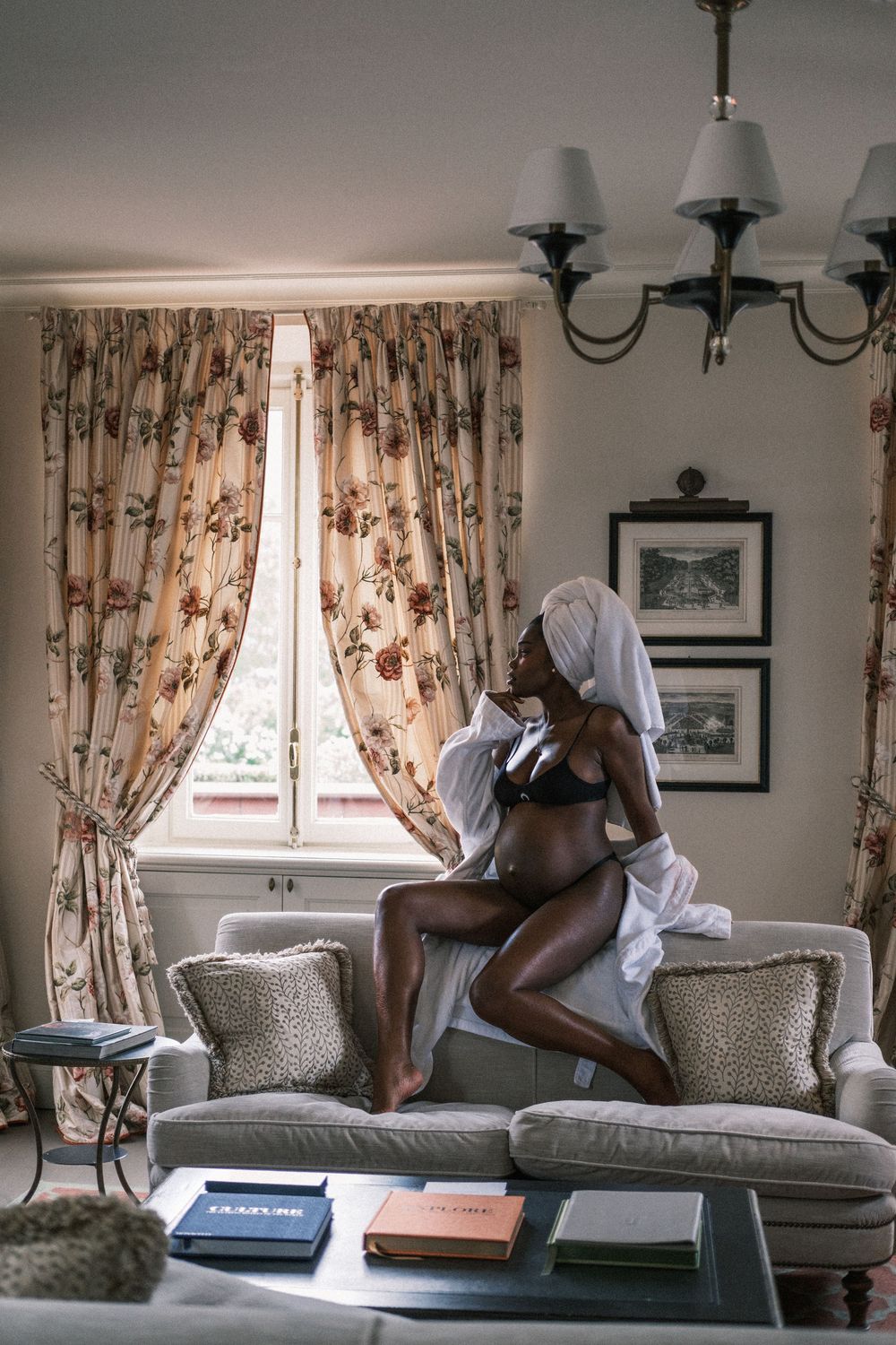 Woman in lingerie and white shirt with towel on head sitting on sofa in elegant living room with floral curtains.