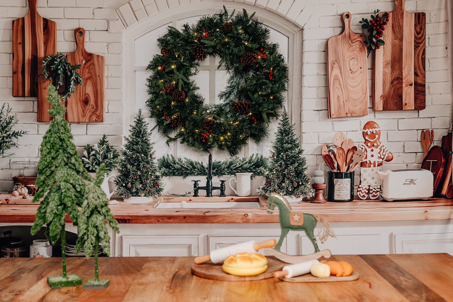 Festive kitchen decorated with Christmas wreath, mini trees, gingerbread man, and wooden cutting boards on white brick wall.