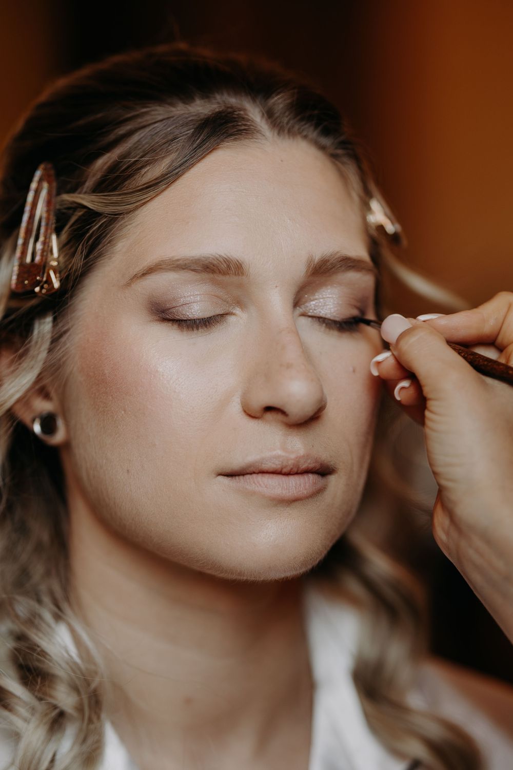 Professional makeup artist applying eyeliner to a blonde bride with hair clips during wedding preparation.