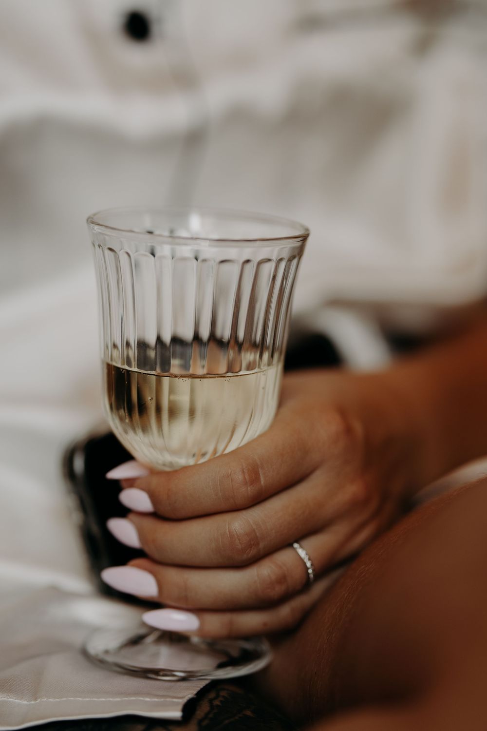Close-up of a hand with pink nails holding a ribbed glass of white wine, wearing a delicate silver ring.