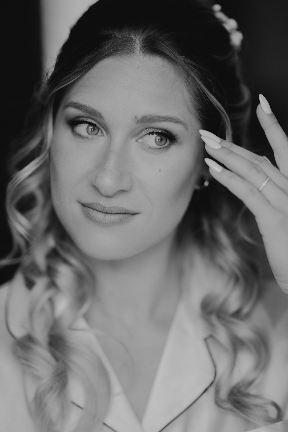 Black and white portrait of a bride with curly hair, floral headpiece, and elegant makeup touching her face gently.