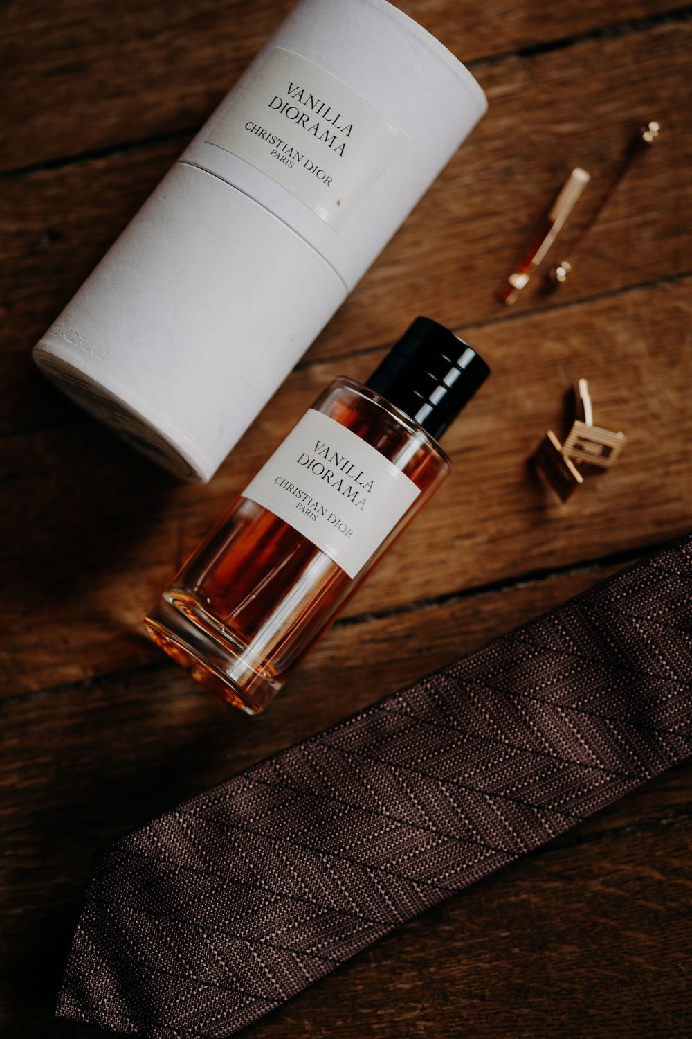 Christian Dior Vanilla Diorama perfume bottle with cufflinks and brown patterned tie on wooden surface.