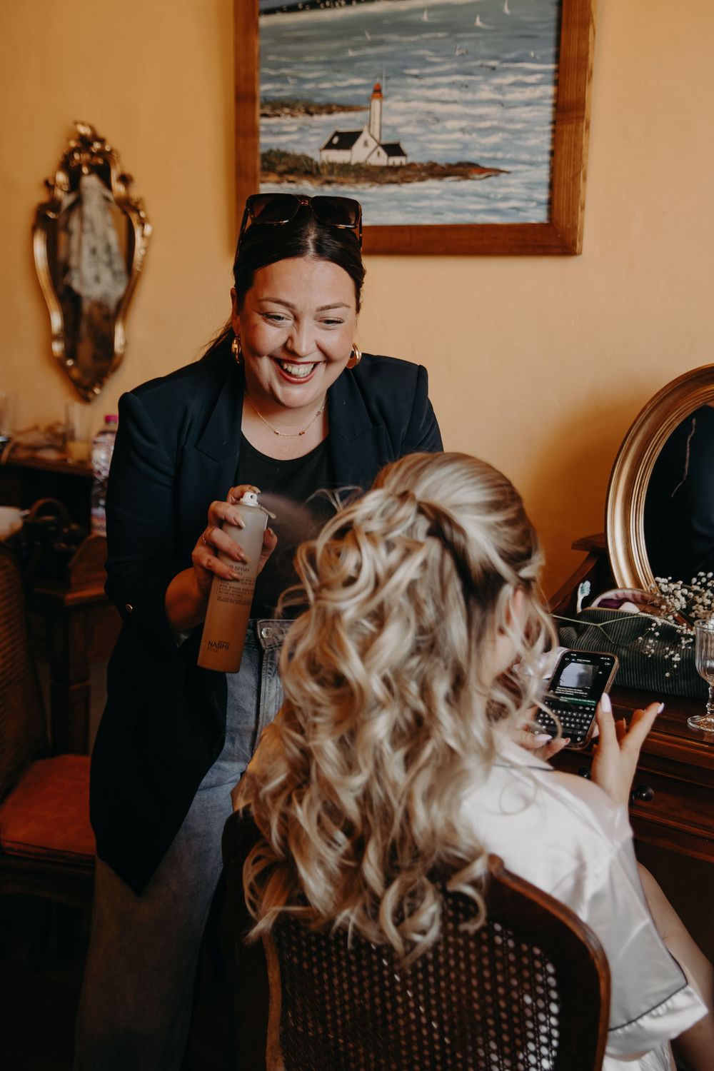 A smiling hairstylist applies hairspray to a bride with elegant curled half-up hairstyle during wedding preparations.