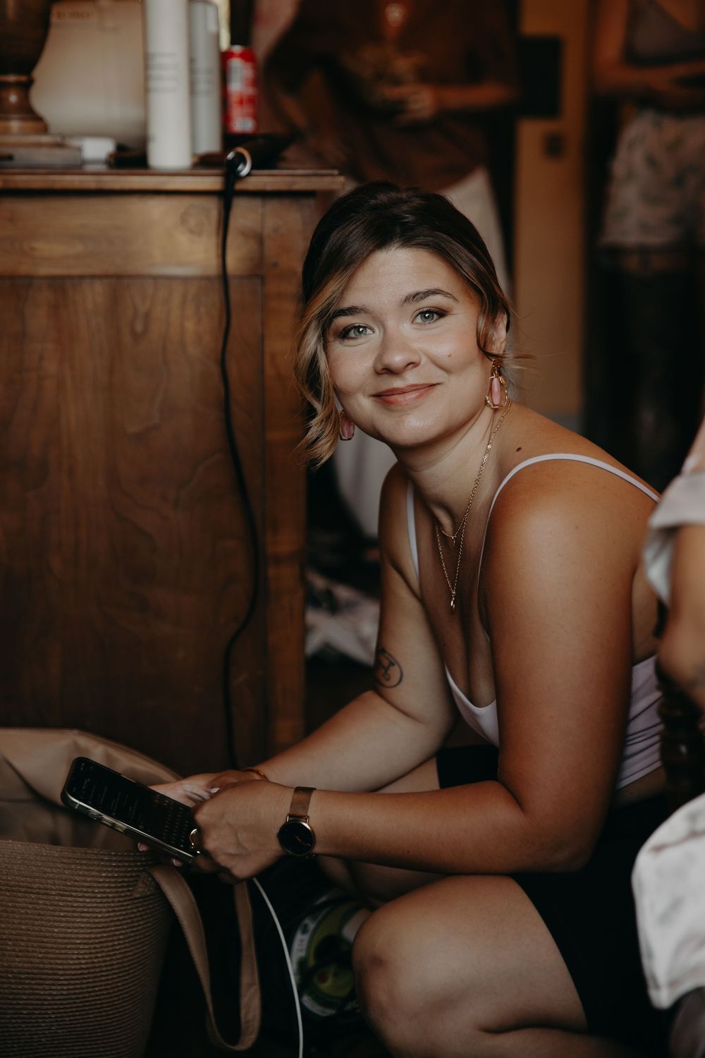 Young woman smiling while holding a phone, wearing a white top and watch, seated near a wooden dresser indoors.