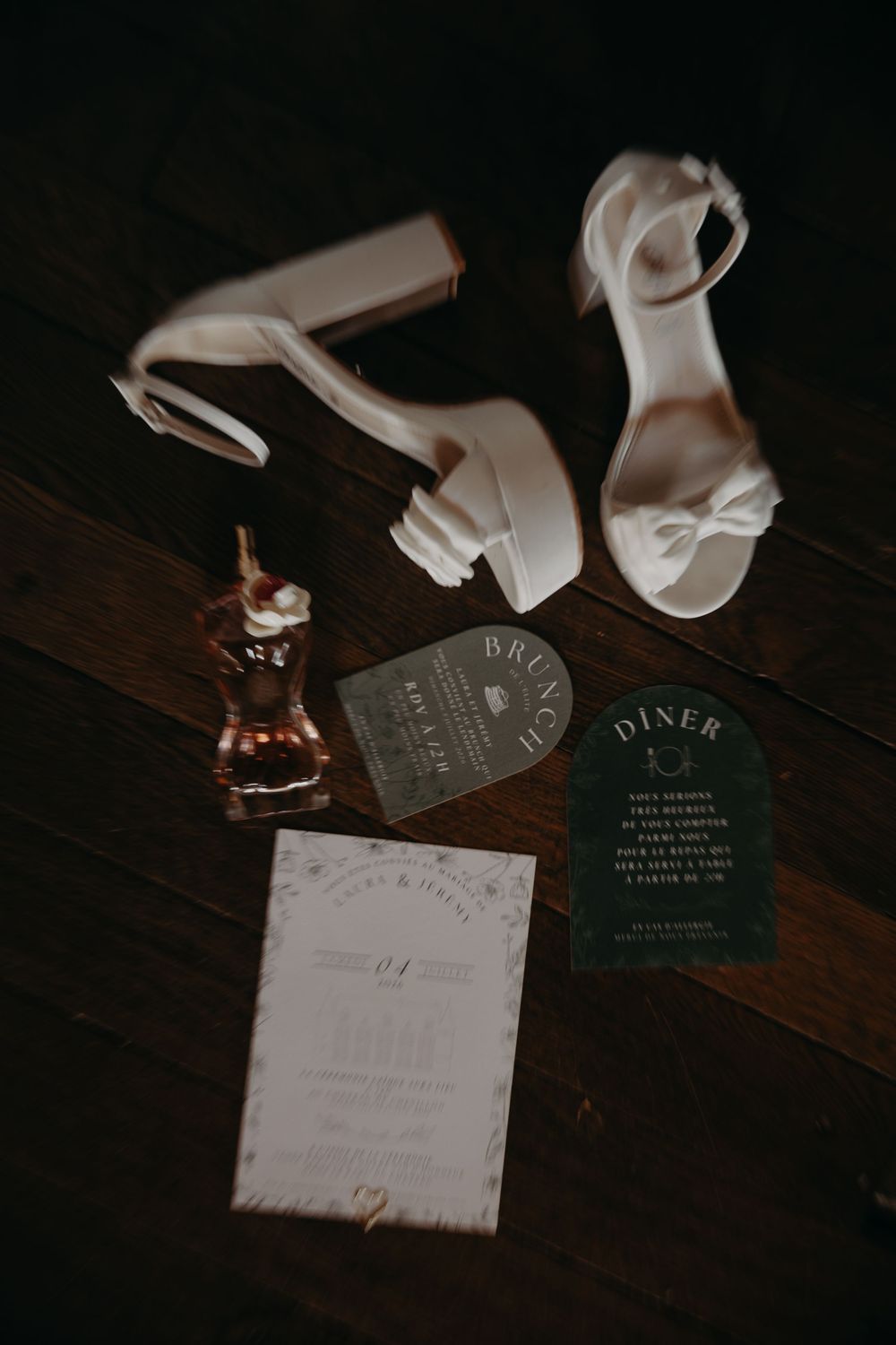 Elegant wedding flat lay featuring white block heels, perfume, green menu cards, and invitation suite on dark wood floor.
