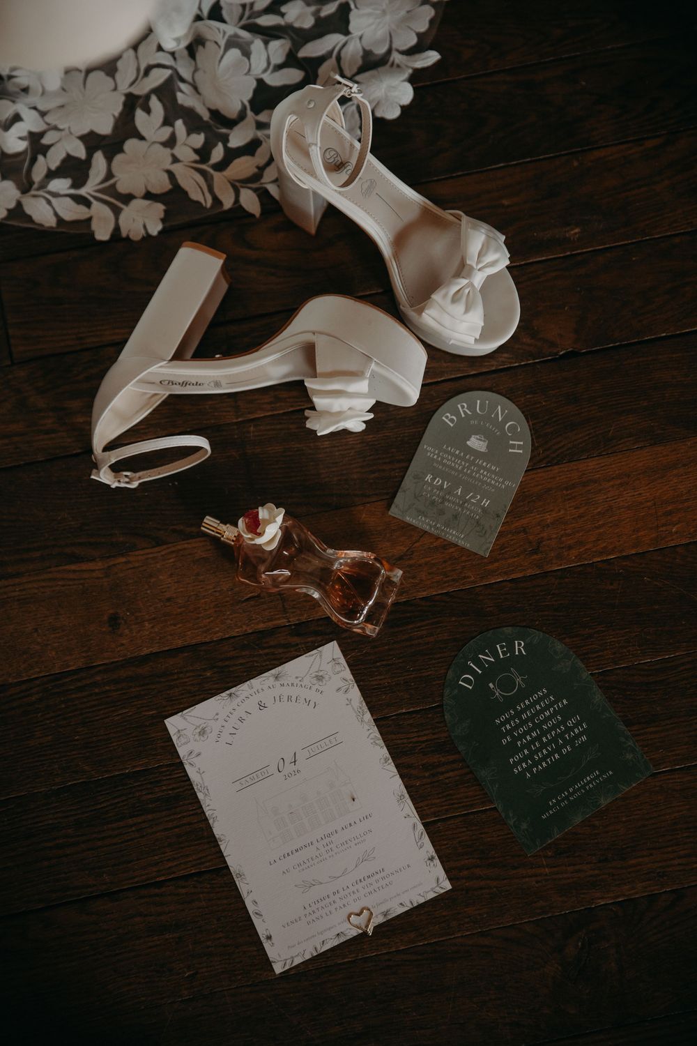 Elegant bridal flat lay featuring white heels, wedding invitations, perfume bottle, and green diner card on dark wood floor.