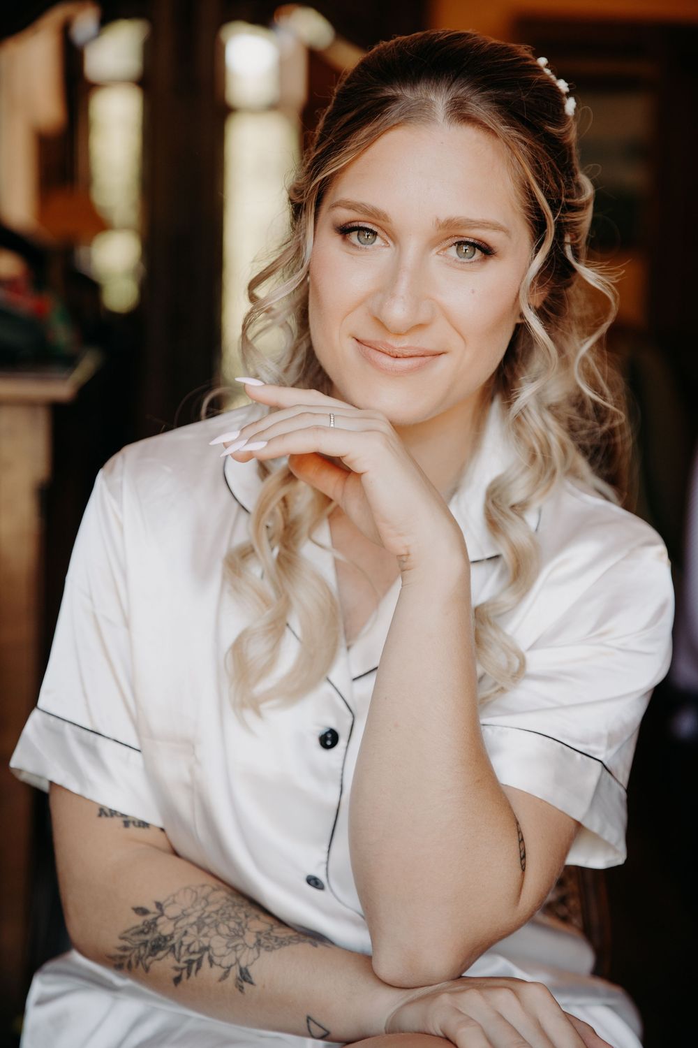 Smiling blonde bride in white satin pajamas with floral arm tattoo and pearl hair accessory posing indoors.