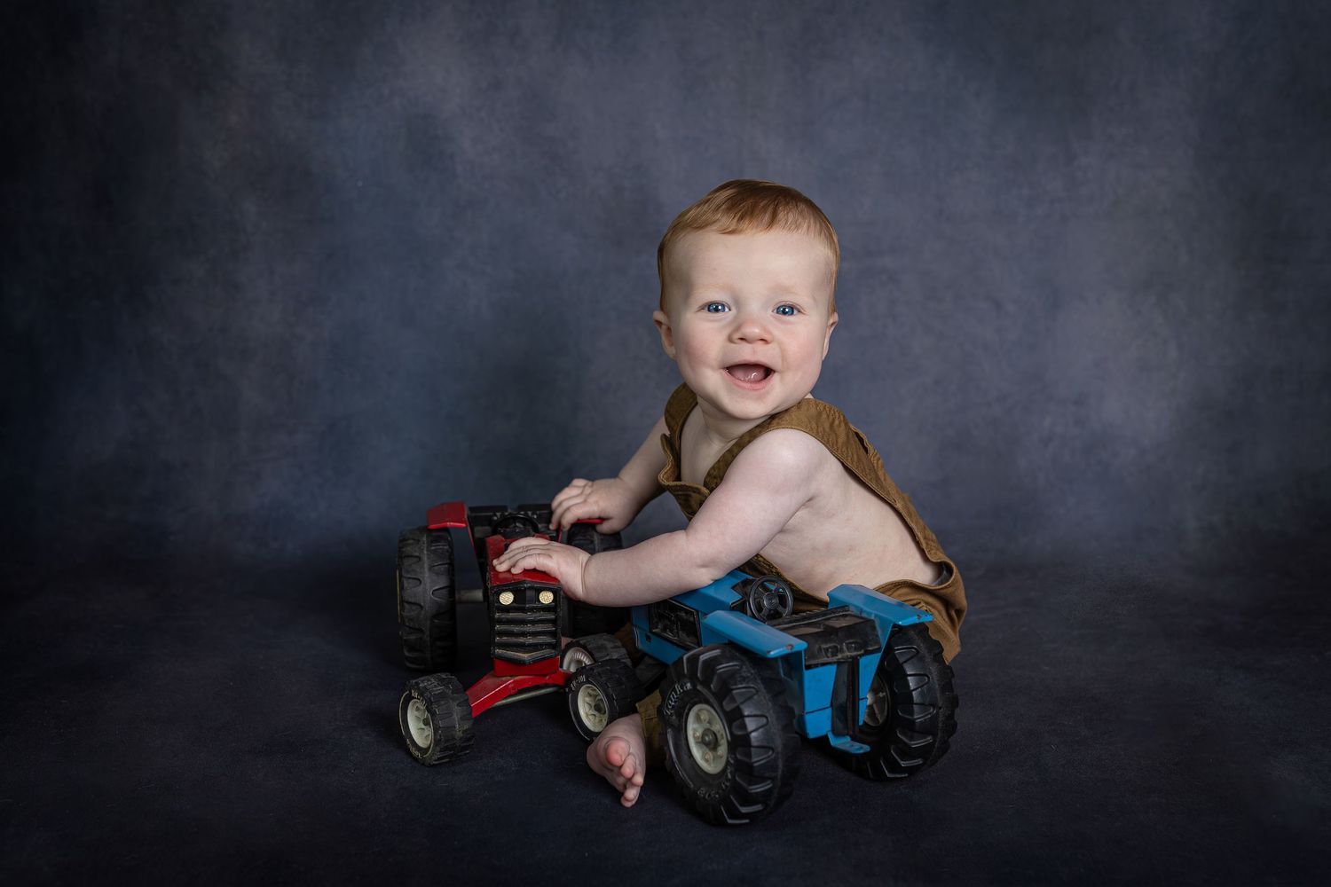 Happy redhead baby boy smiles while playing with toy tractors against gray studio backdrop.