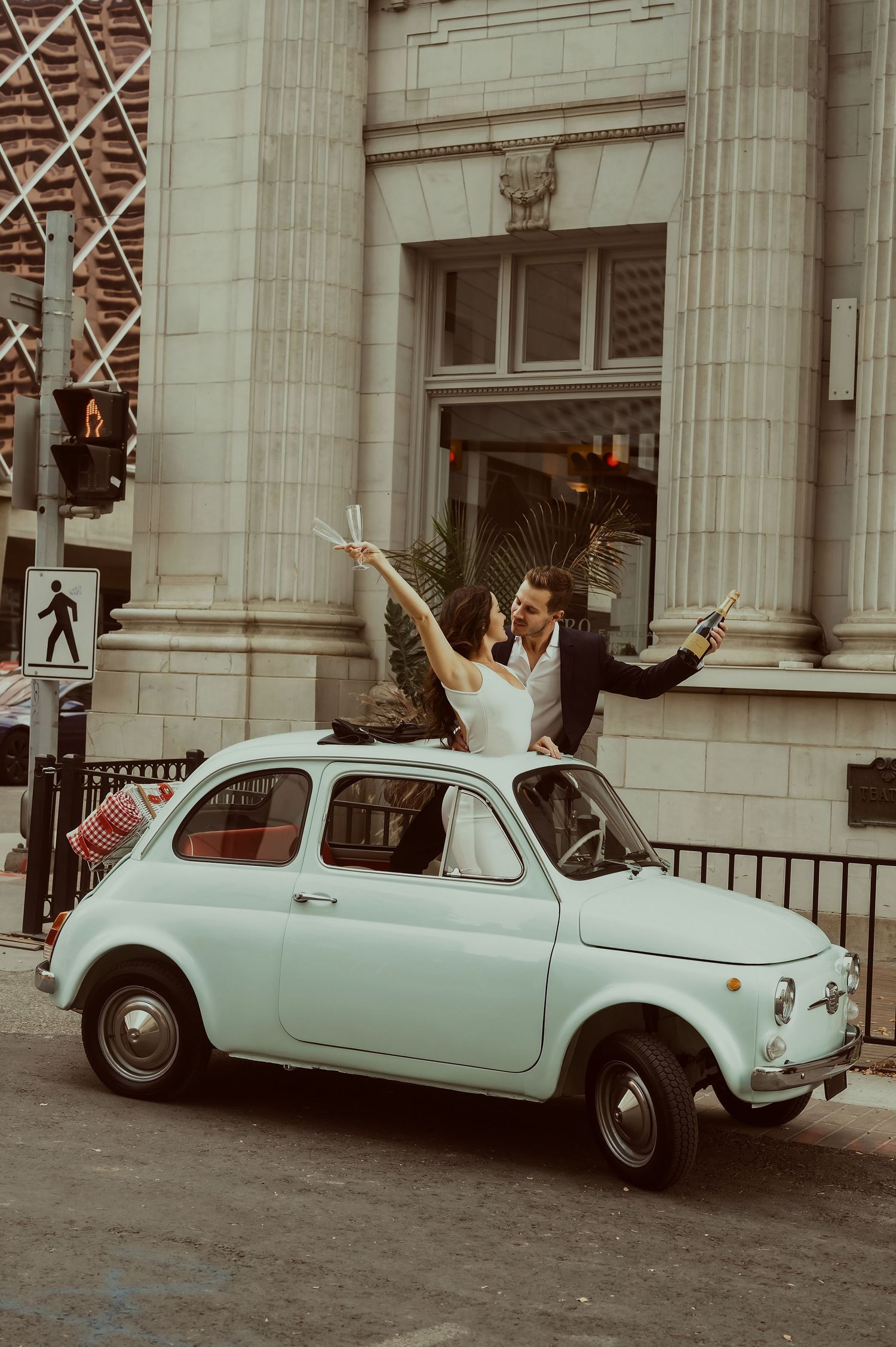 Couple celebrating on a vintage mint green Fiat 500 car on a city street, raising champagne bottles joyfully.