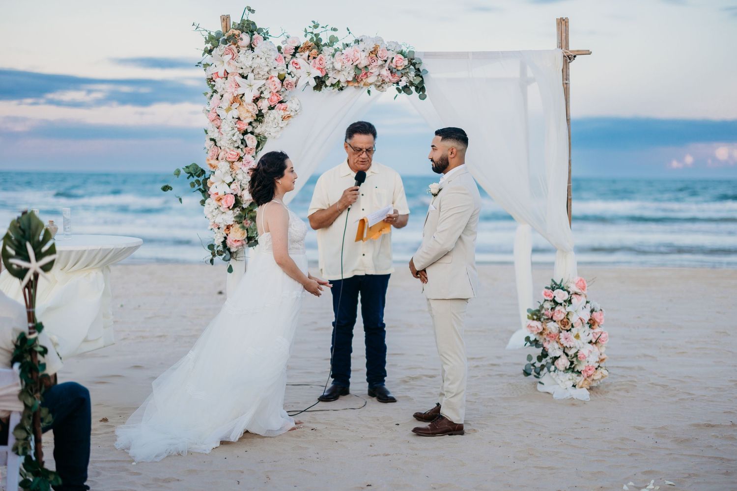 Couple exchanging vows at a romantic beach wedding ceremony under a floral arch at sunset.