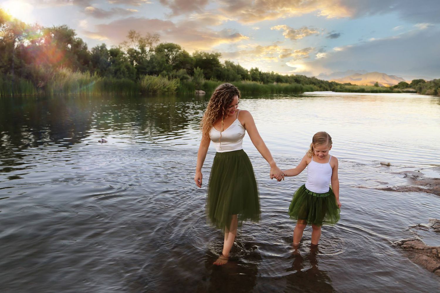 Mother and young daughter in matching green tulle skirts hold hands while wading in a calm river at sunset.
