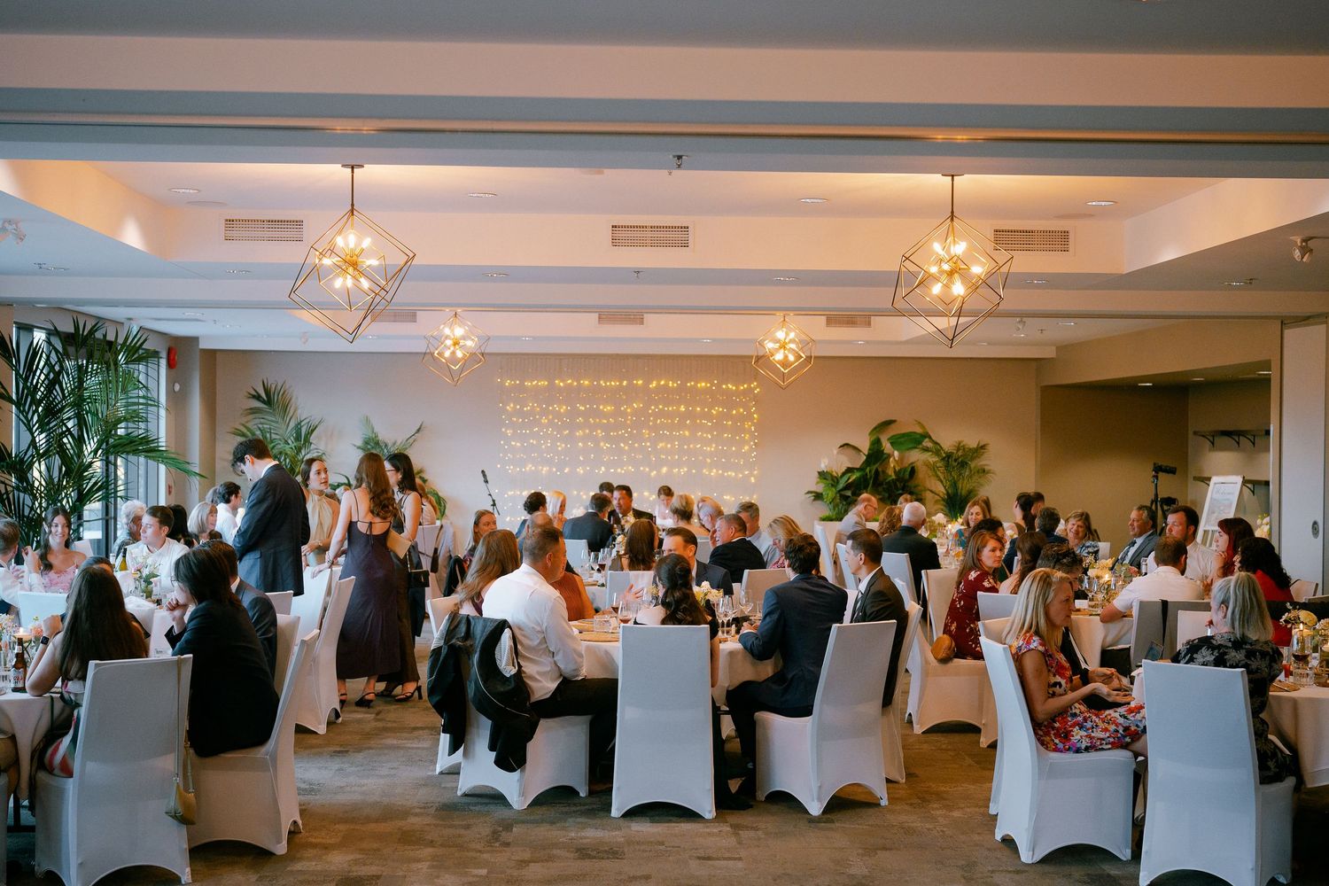 Elegant wedding reception with guests seated at round tables with white chairs in a warmly lit ballroom.