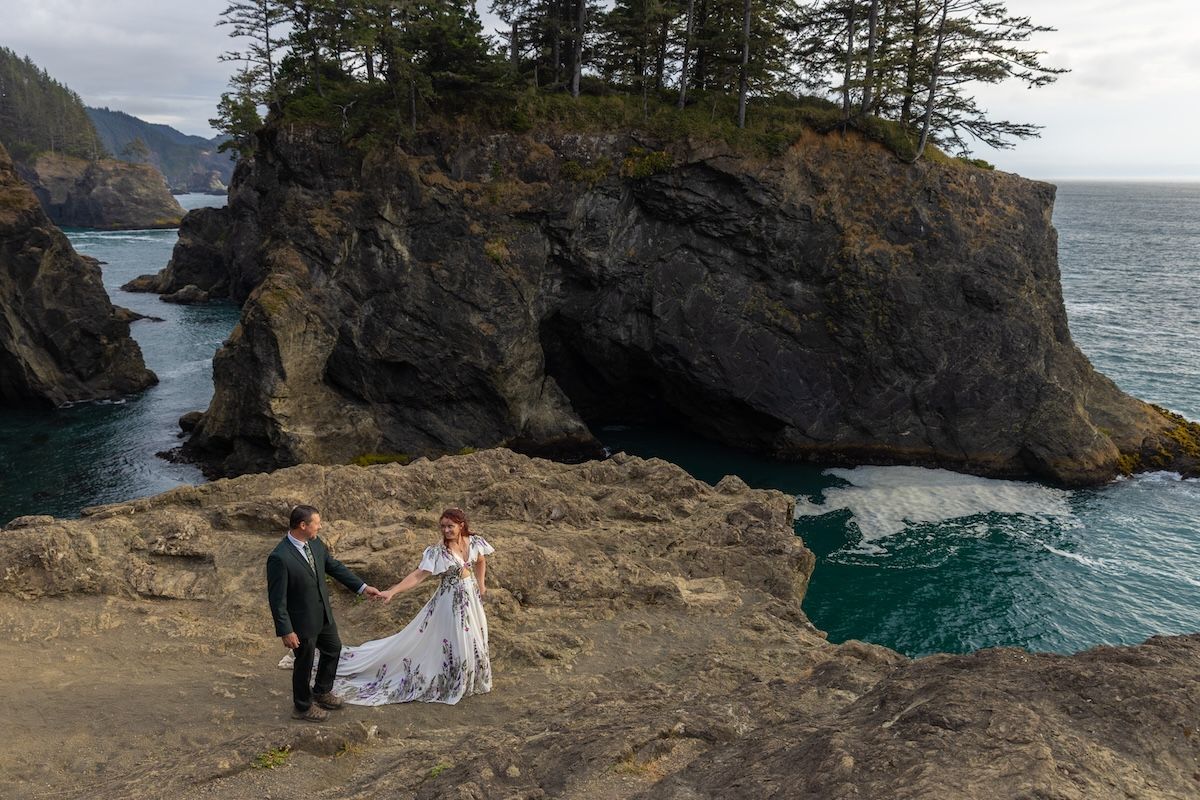 Couple in wedding attire walks along a scenic Pacific Northwest beach with dramatic sea stacks and turquoise water.