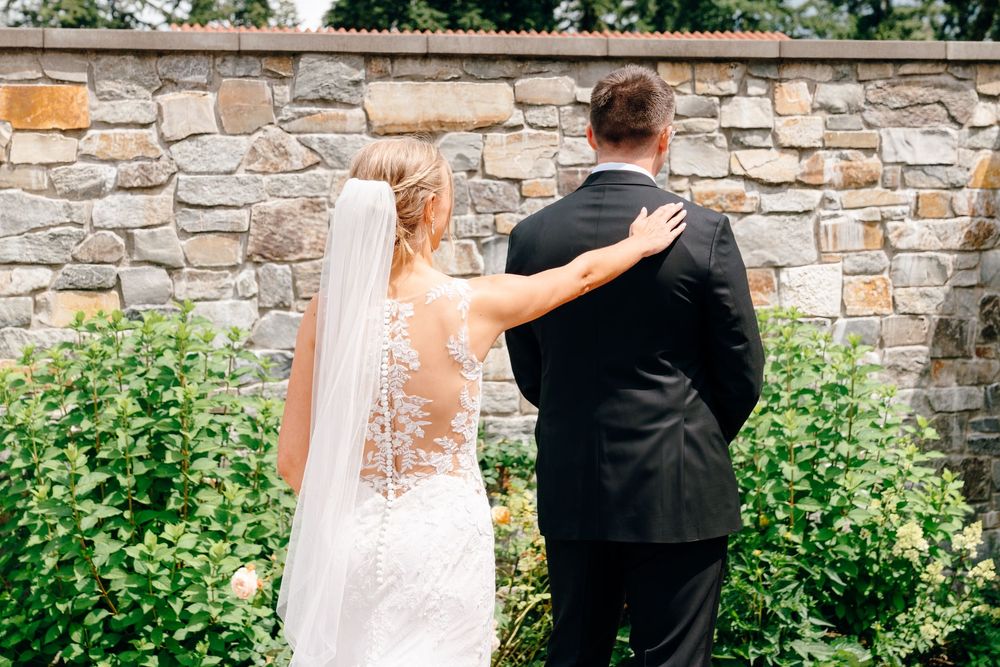 Bride and groom seen from behind in garden, bride wearing lace gown with veil, groom in black tuxedo.