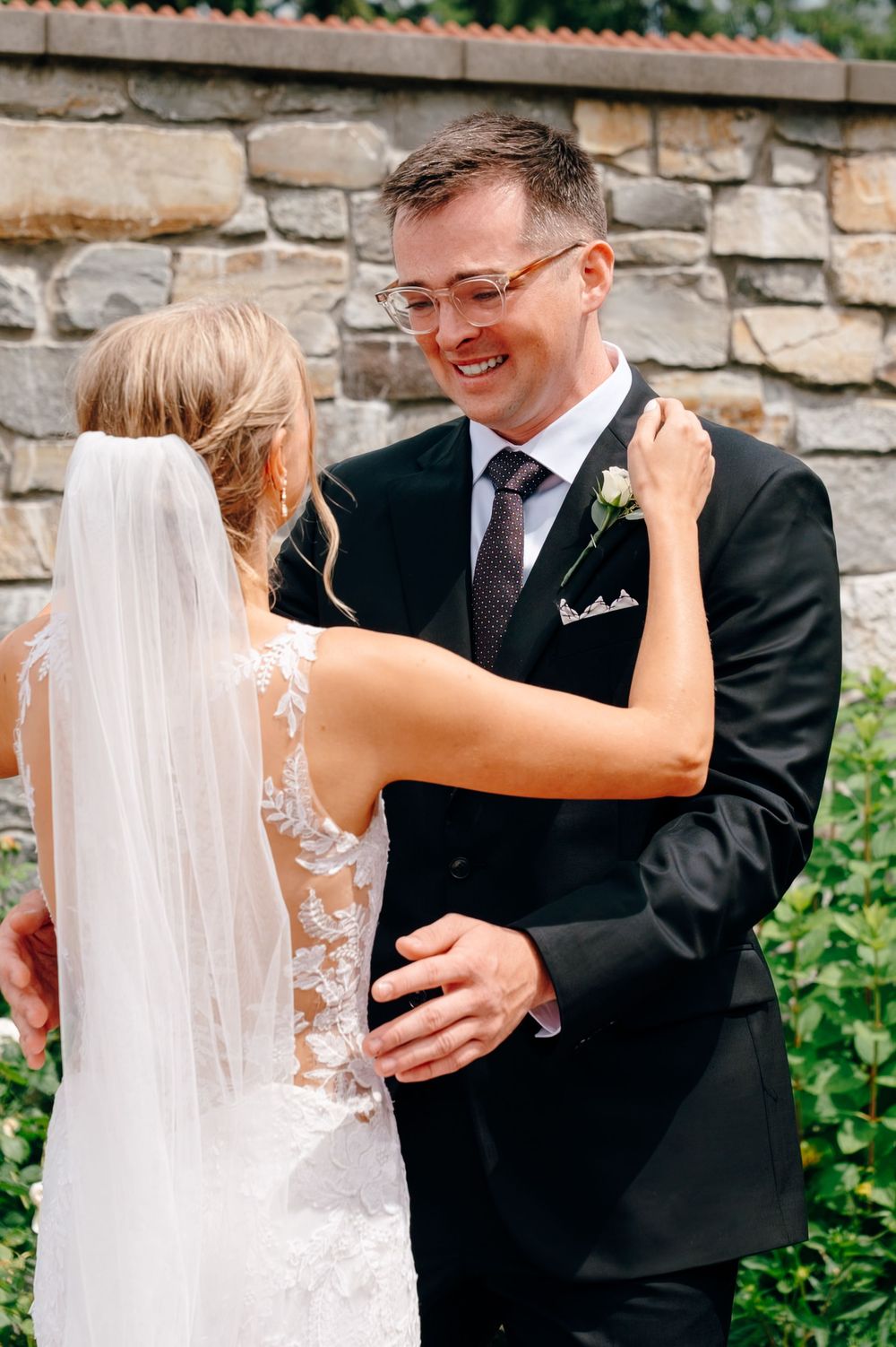 Groom in black suit smiling joyfully as bride in lace wedding dress and veil embraces him outdoors.