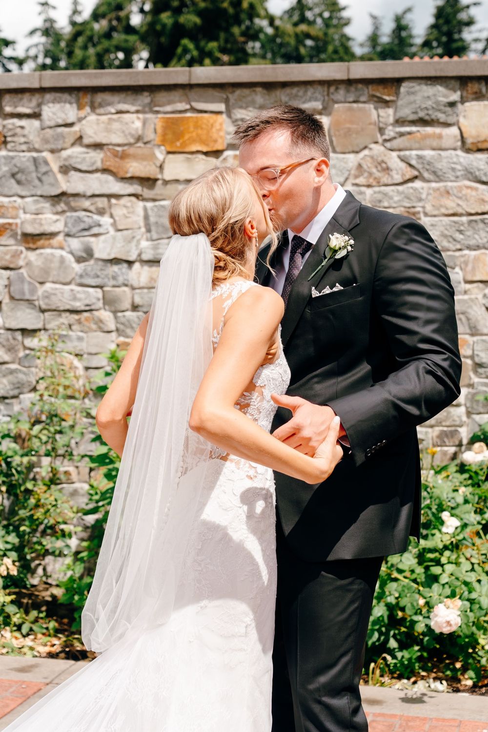 Bride and groom sharing a romantic kiss outdoors, with a stone wall and garden in the background.