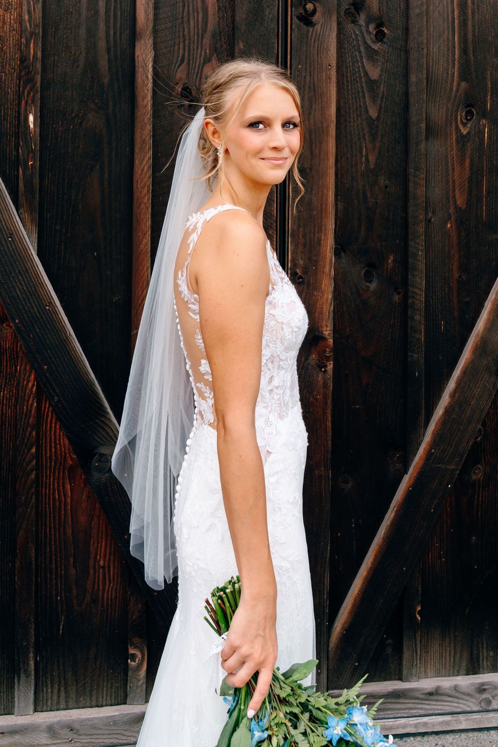 Bride in white lace gown and veil holds blue bouquet, posing in front of rustic dark wood barn doors.