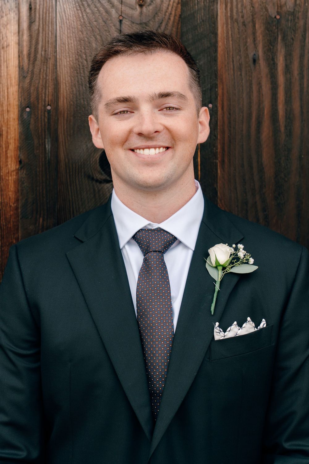 Smiling groom in dark suit with boutonniere and pocket square poses against wooden backdrop.
