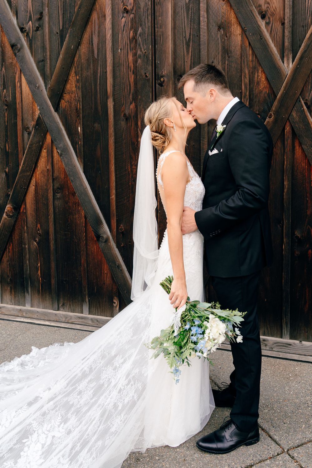 Bride and groom share a kiss in front of rustic wooden barn doors, bride holding blue and white bouquet.