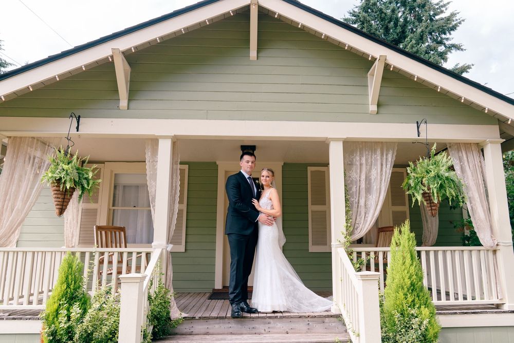 Bride and groom posing on porch of charming green cottage wedding venue with hanging ferns and white curtains.