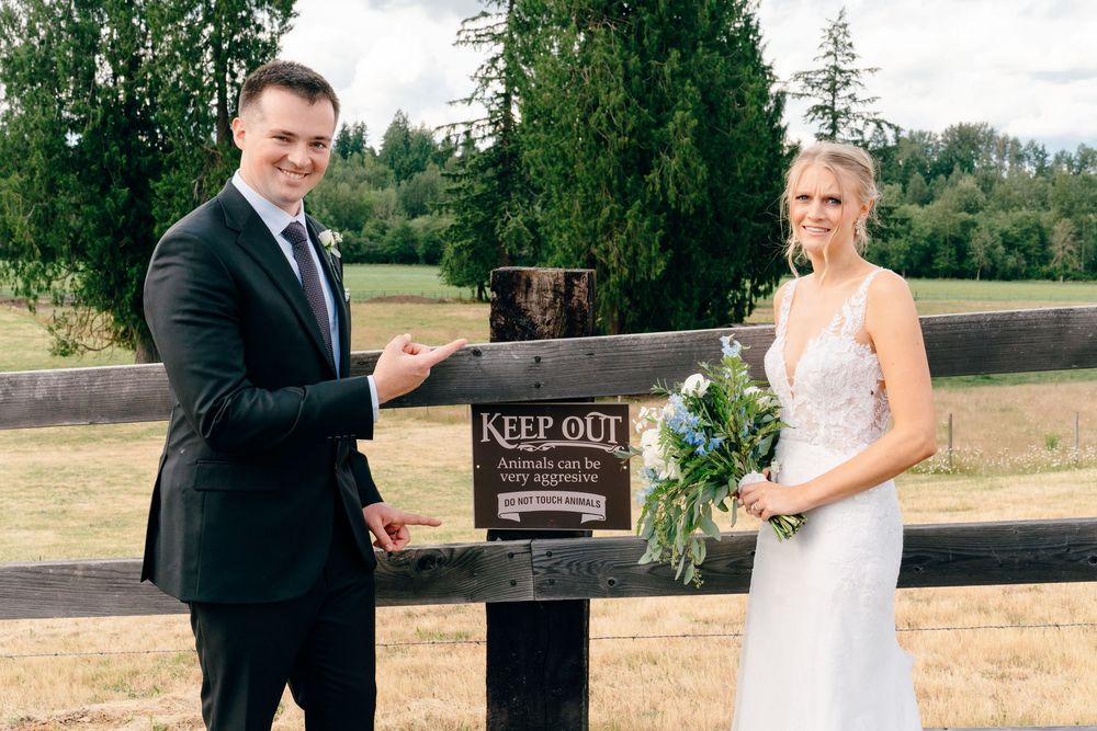 Bride and groom posing beside a Keep Out sign outdoors at a wedding venue surrounded by green trees.
