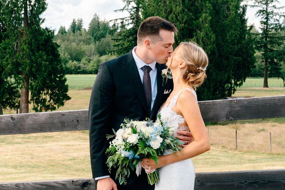 A bride and groom share a romantic kiss outdoors at a wedding venue, with the bride holding a white and blue bouquet.