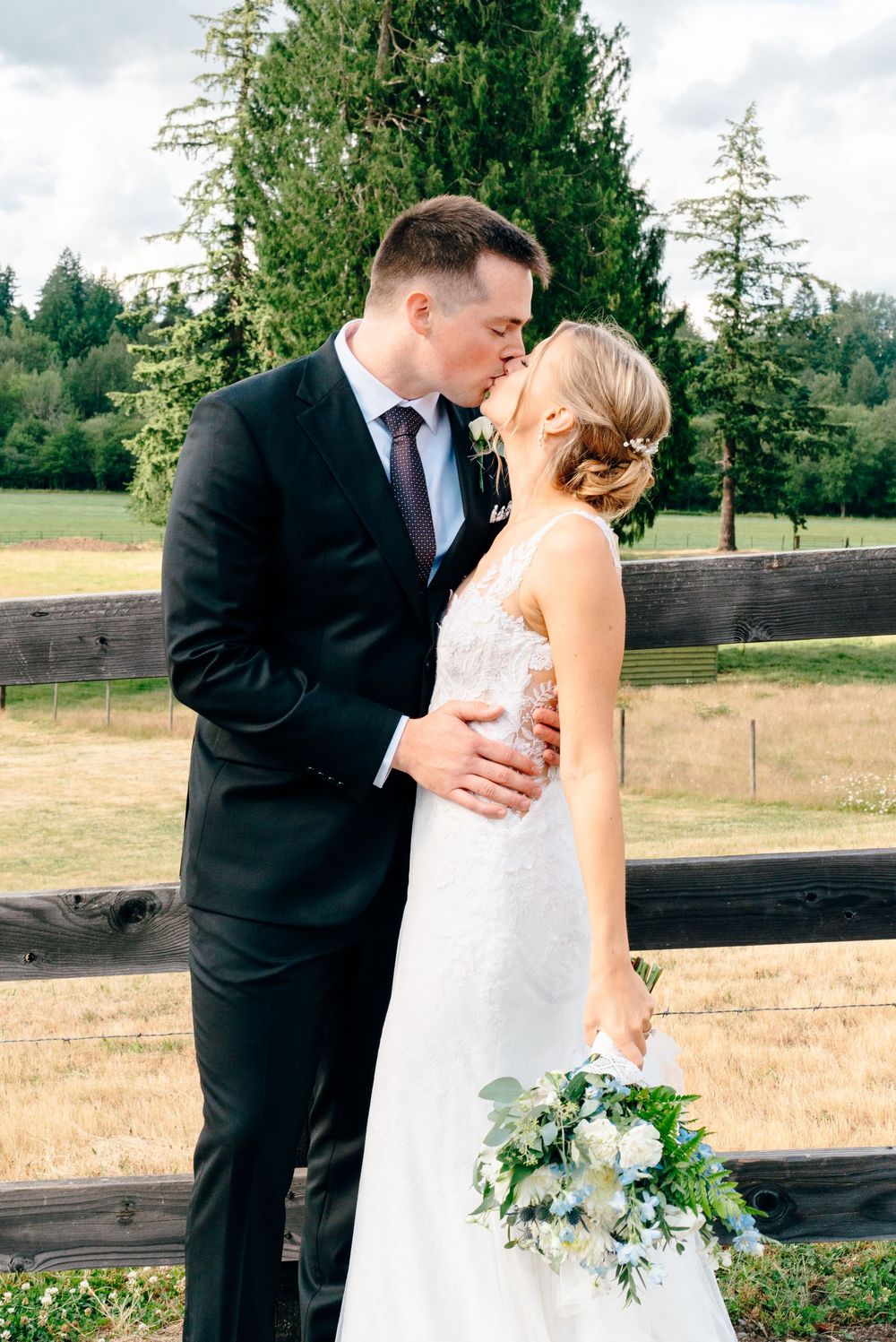 A bride and groom share a kiss outdoors near a wooden fence, surrounded by lush green trees on their wedding day.