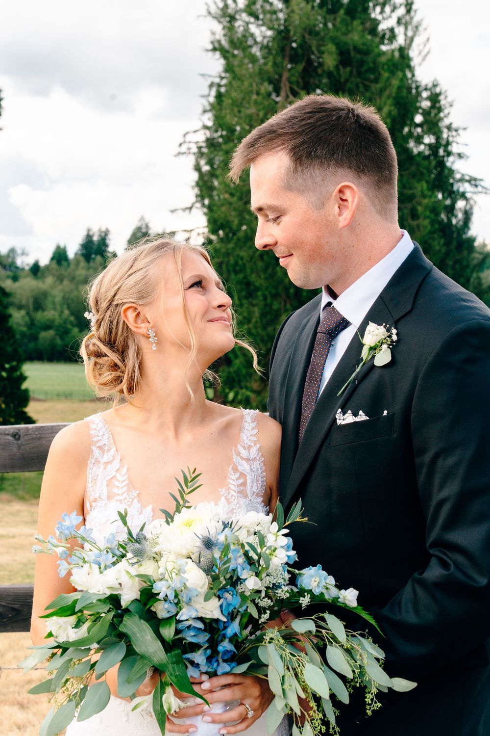Bride and groom gazing at each other outdoors, bride holding blue and white floral bouquet, groom in dark suit with boutonniere.