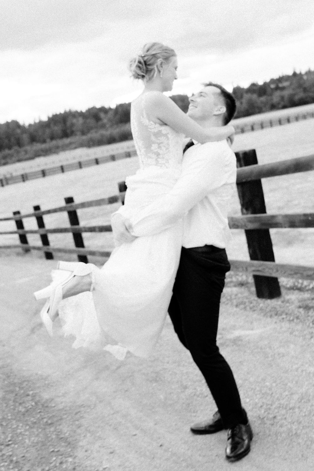 Groom lifting bride in wedding attire outdoors by rustic fence in black and white photo.