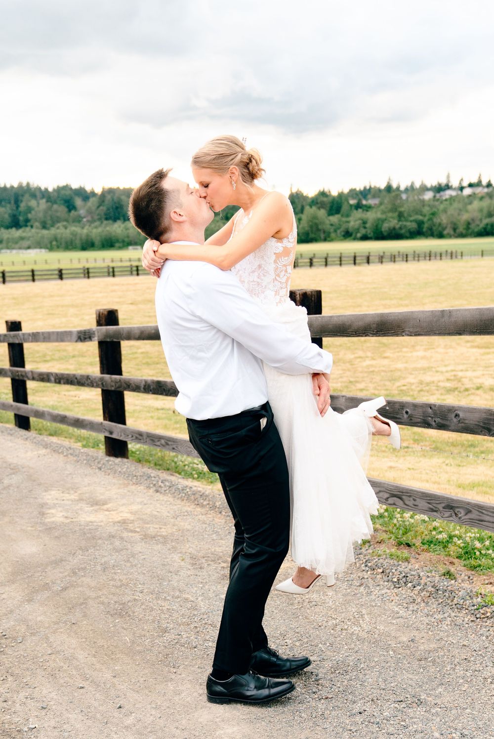Groom lifts bride in white wedding dress for a romantic kiss outdoors by a wooden fence at a farm venue.