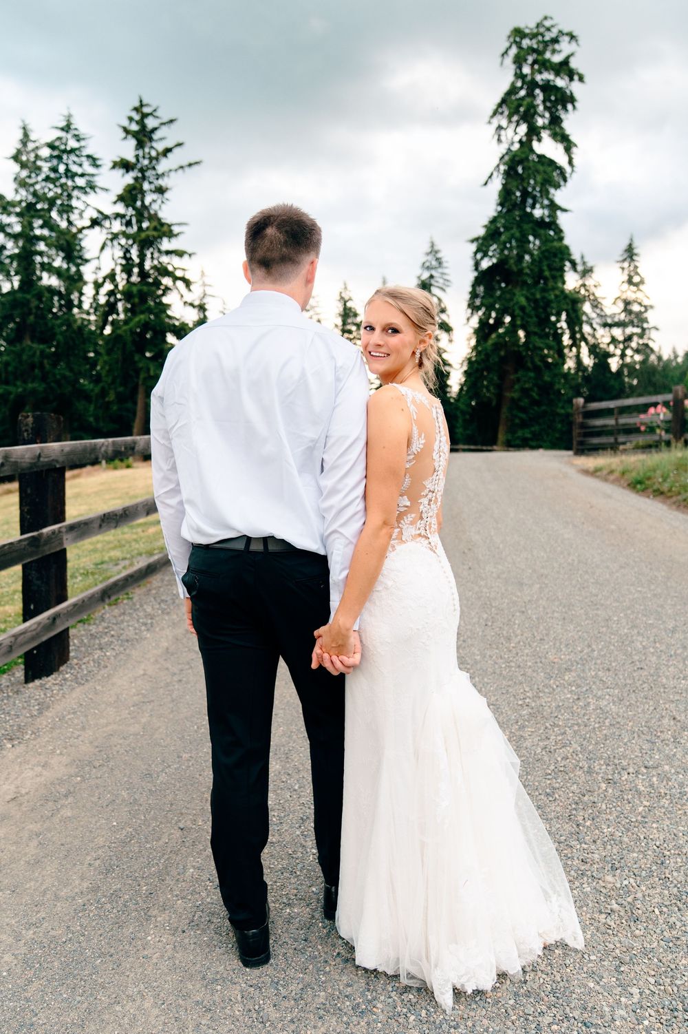 Bride in lace backless gown and groom in white shirt walk hand-in-hand down a tree-lined gravel path.