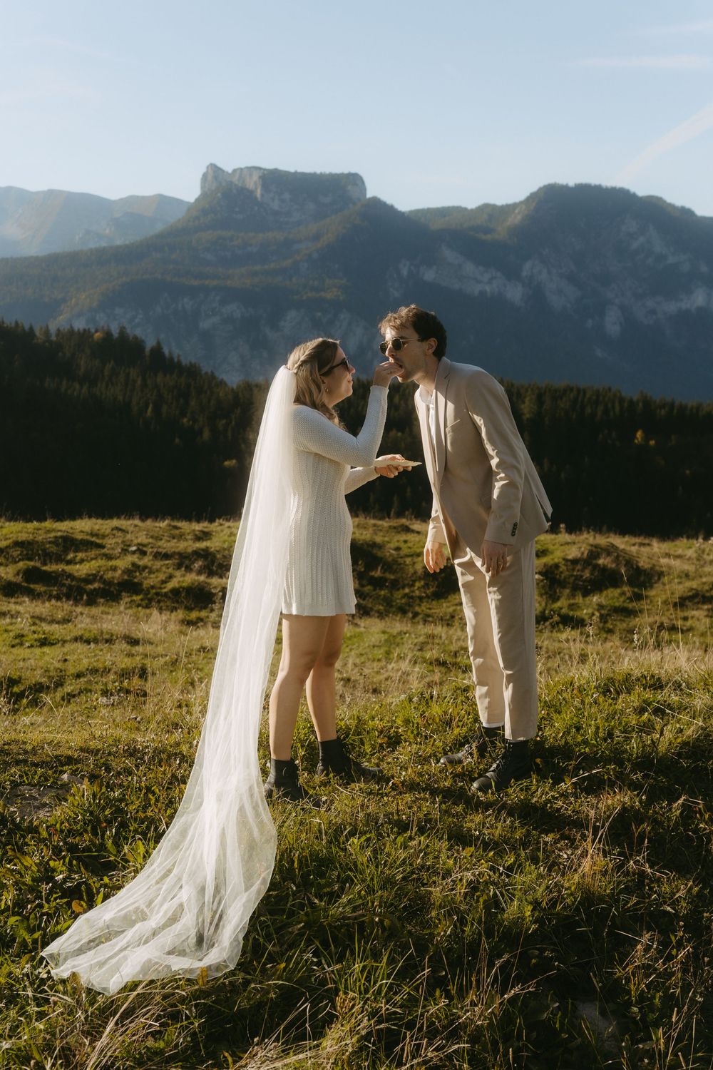 Bride and groom face each other smiling in sunny alpine meadow with rocky mountain backdrop.