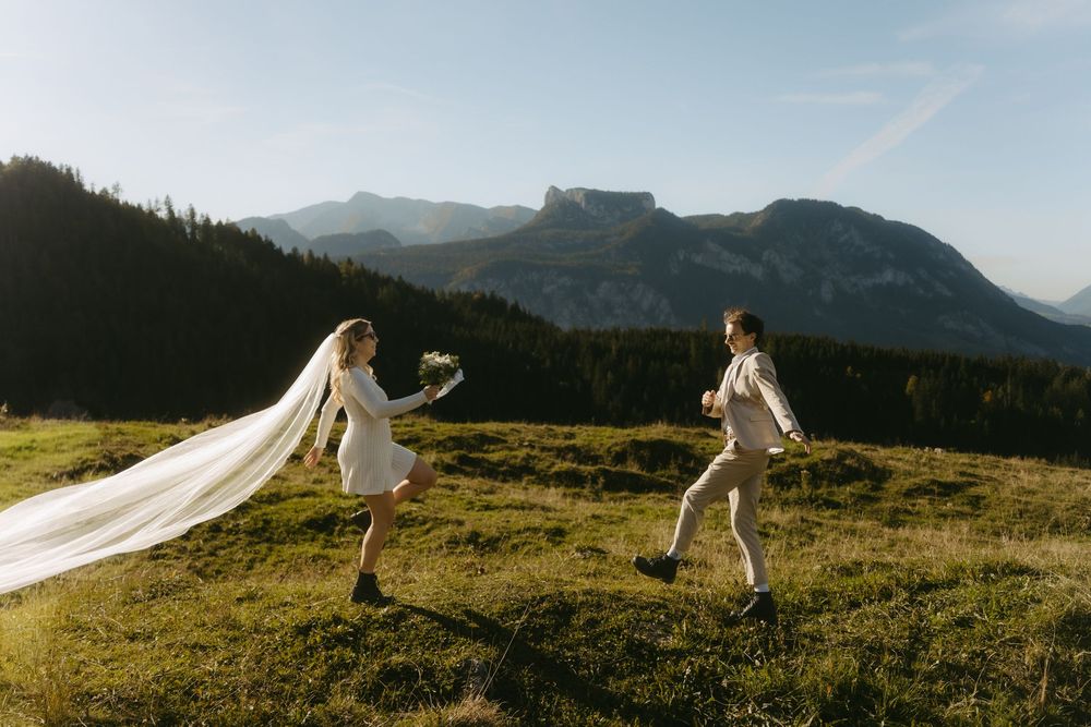 Bride and groom running on alpine meadow with dramatic mountain backdrop and flowing wedding veil in golden sunlight.