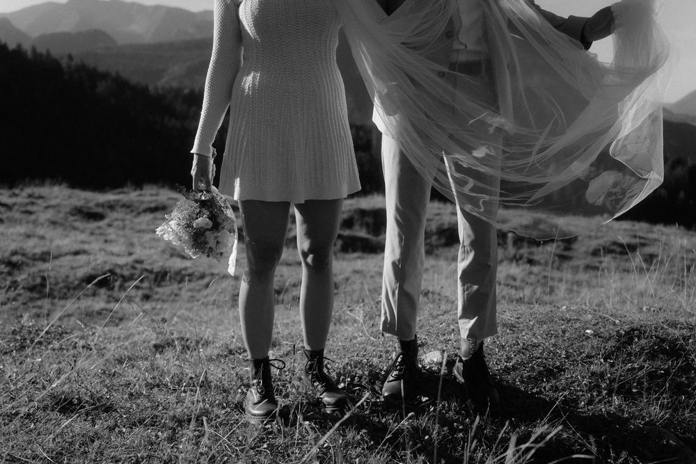 Black and white photo of bride and groom standing on mountain meadow, bride holding bouquet with veil blowing in wind.