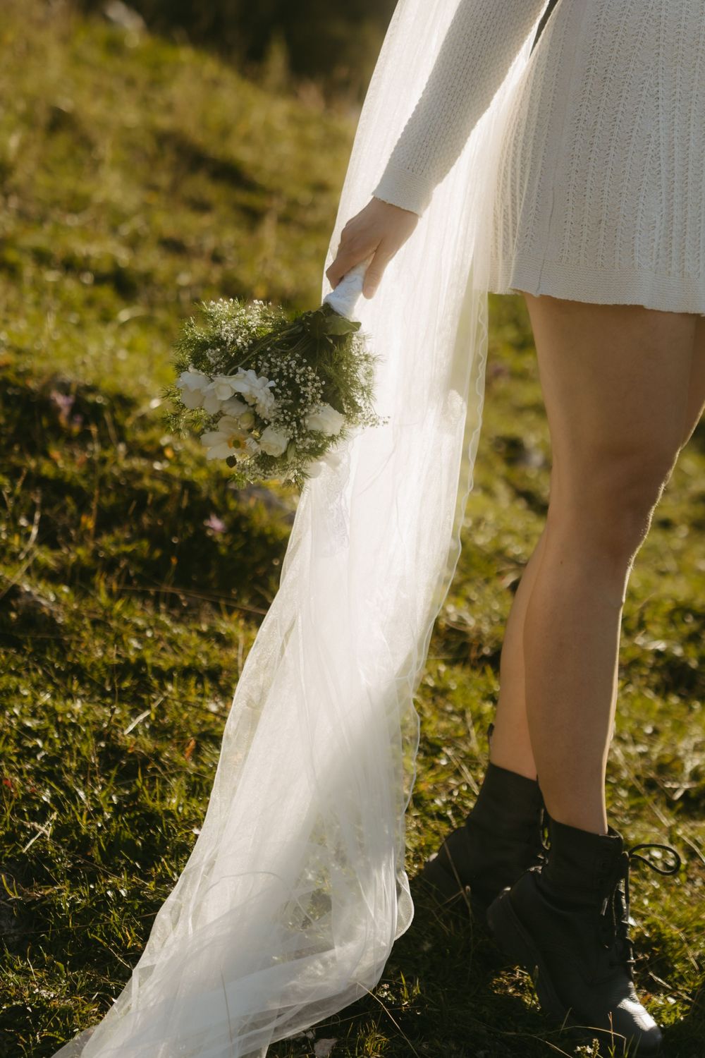 Bride in white knit dress holds bouquet of white flowers with long veil trailing on grass.