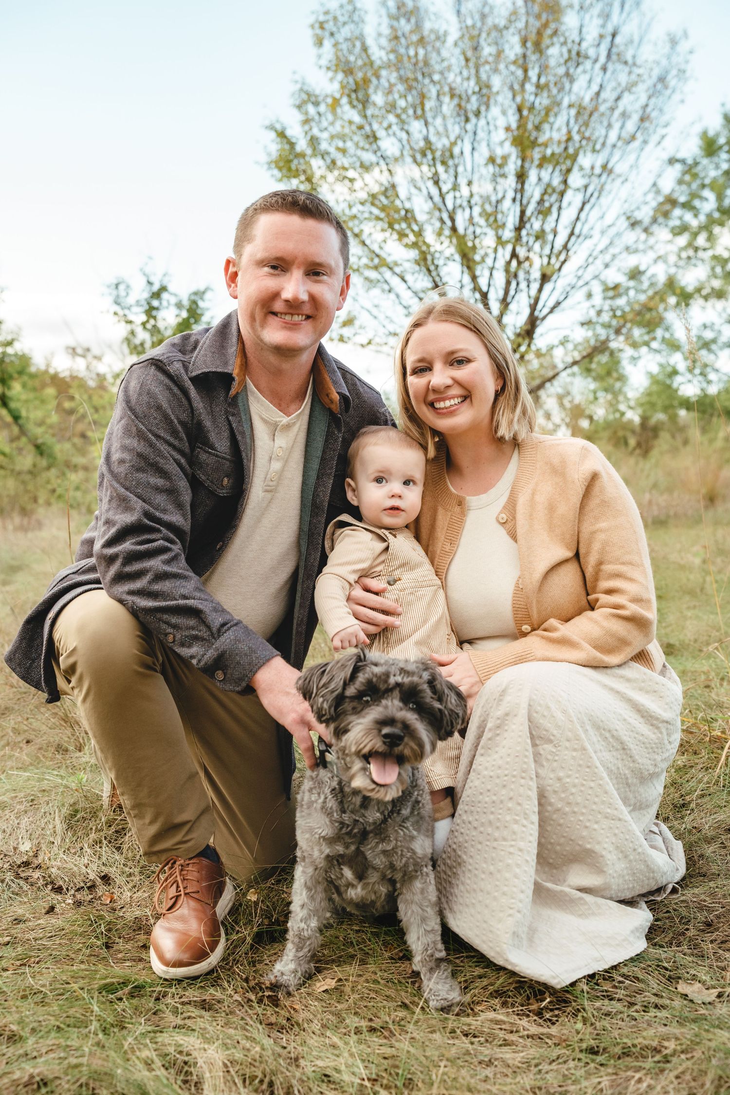 A happy young family with a baby boy poses outdoors in a golden meadow during a fall family portrait session.