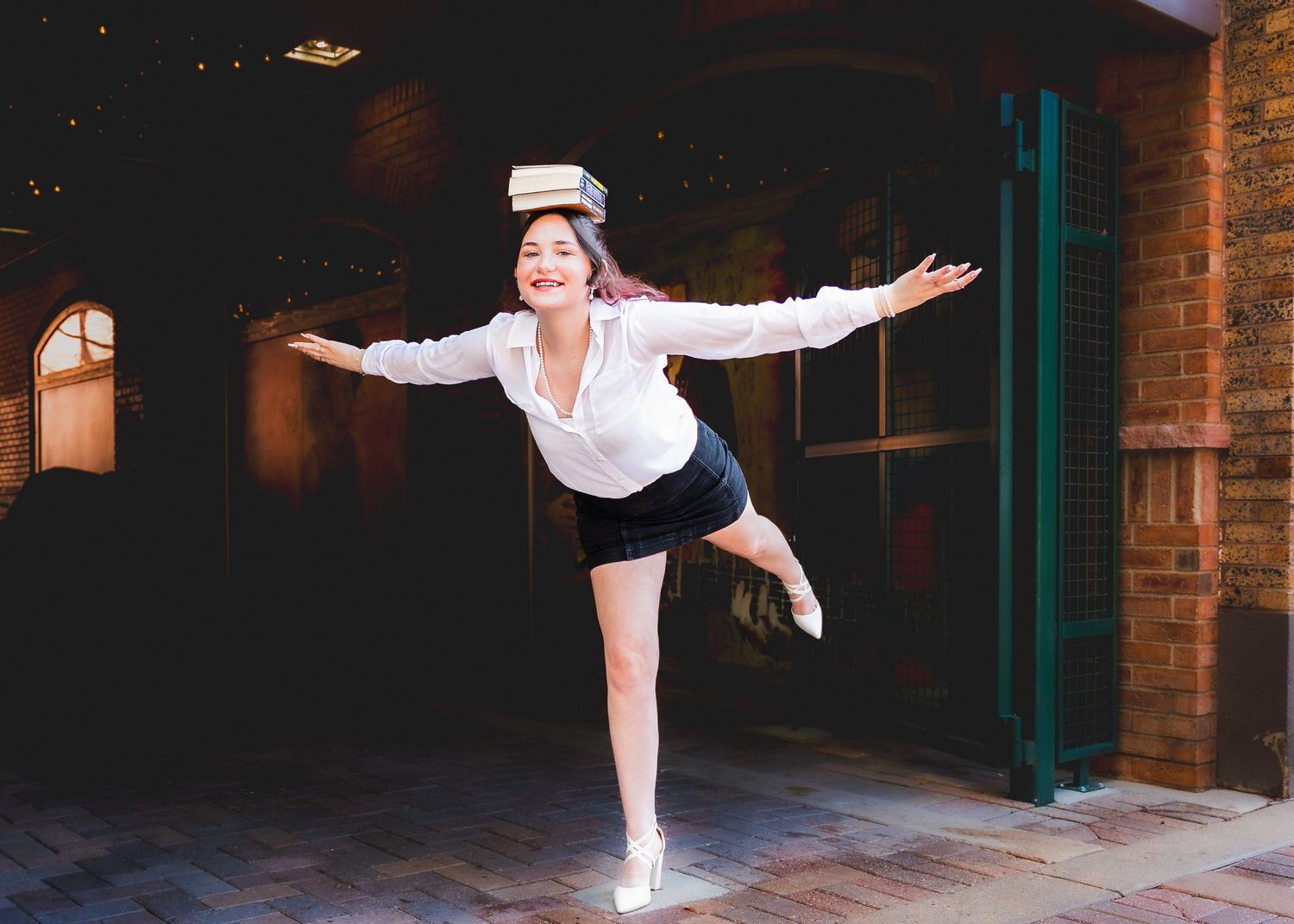Young woman balancing books on her head while posing on one leg in a white shirt and black shorts.