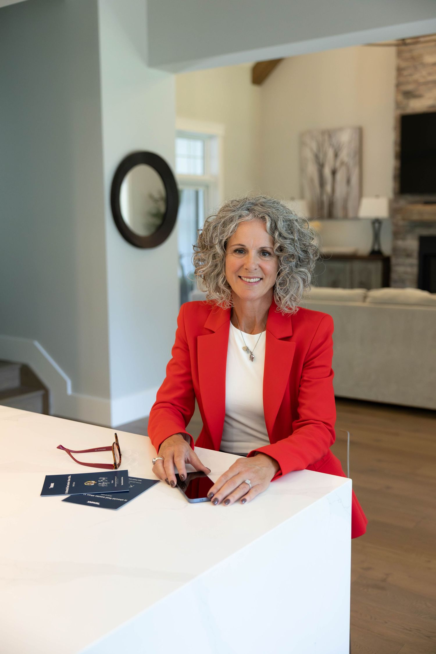 A smiling woman with curly gray hair in a red blazer sits at a white counter in an elegant home interior.